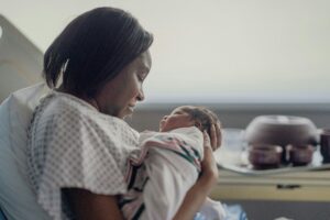 A mother in a hospital gown sits in a hospital bed, cradling her newborn baby wrapped in a striped hospital blanket. She gazes at the baby with warmth and love, while soft light fills the room.